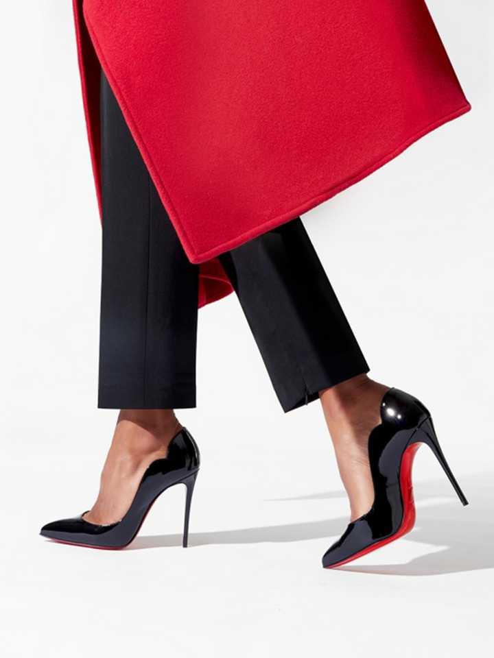 Person wearing black high-heeled shoes with red soles, black pants, and a red coat on a white background.