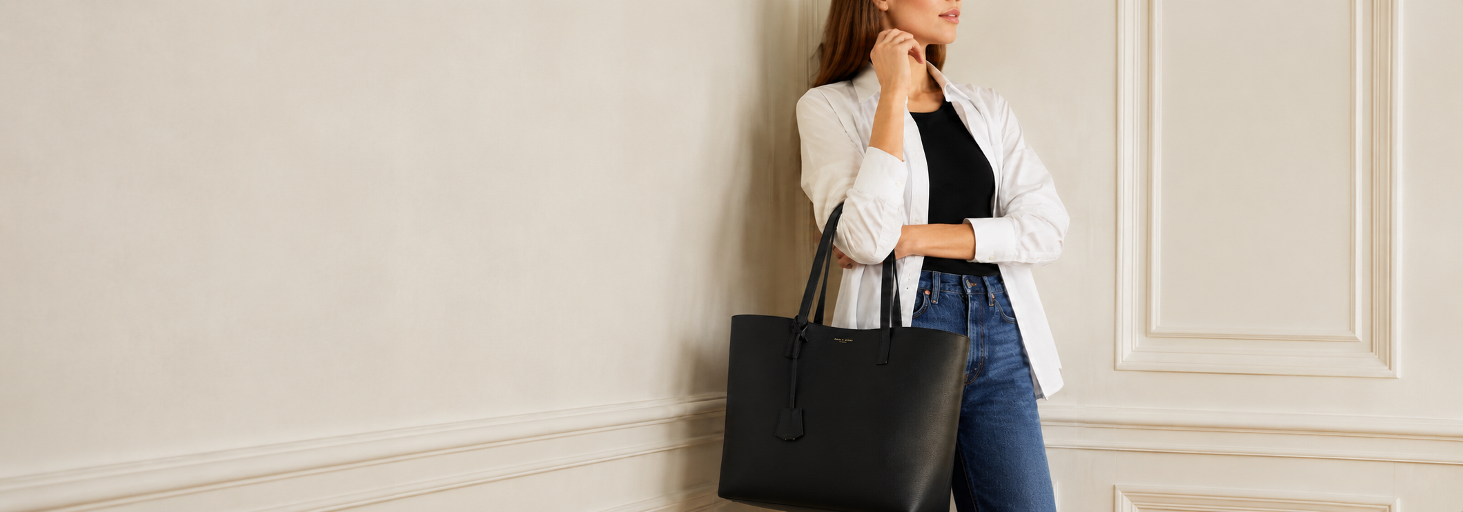 A woman in a white blazer, black top, and blue jeans stands against a cream-colored wall, holding a large black leather tote bag.