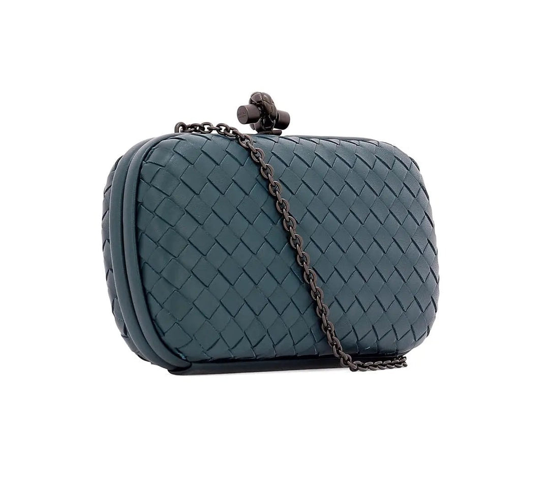 A woven leather clutch bag in a muted teal blue-green color, featuring a dark metal chain strap and a decorative clasp closure, set against a plain white background.